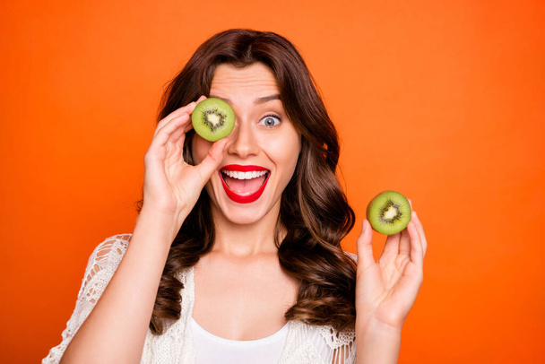 Photo of cheerful cute nice sweet pretty curly wavy girlfriend looking out of half of kiwi expressing positive emotions on face isolated over vivid orange color background - Fotoğraf, Görsel
