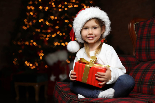 Cute little child with Christmas gift sitting in armchair at home - Foto, Imagen