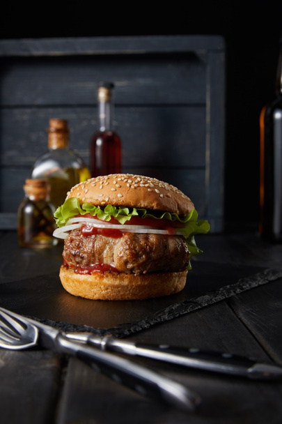 selective focus of burger on dark chopping board near fork, knife, oil and vinegar bottles isolated on black - Photo, Image