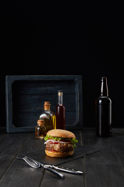 burger na ciemnej drewnianej powierzchni, widelec, nóż i butelki oleju izolowane na czarno - Zdjęcie, obraz