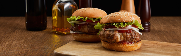 panoramisch shot van twee smakelijke hamburgers op houten plank, geoogste olieflessen, zout en peper molens geïsoleerd op zwart - Foto, afbeelding