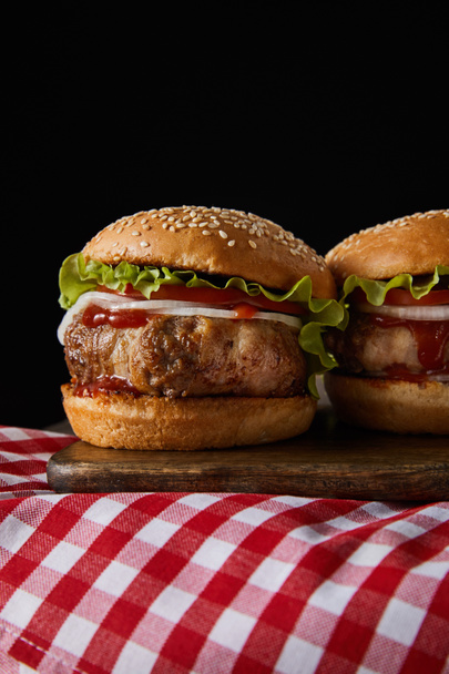 two burgers on wooden chopping board on checkered tablecloth isolated on black - Photo, Image
