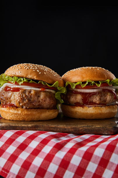two hamburgers on wooden chopping board on checkered tablecloth isolated on black - Fotoğraf, Görsel