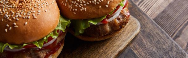 panoramatický vysokoúhlý záběr hamburgerů na dřevěné řezací desce, příbory, olejové, octové a pivní láhve  - Fotografie, Obrázek