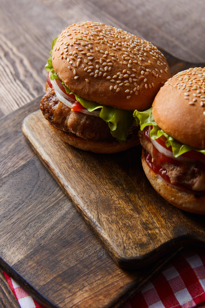 high angle view of two burgers on wooden cutting board on checkered tablecloth - Photo, Image