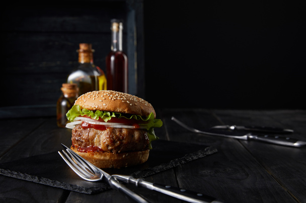 selective focus of burger on dark surface, vinegar and oil bottles isolated on black - Foto, Imagem