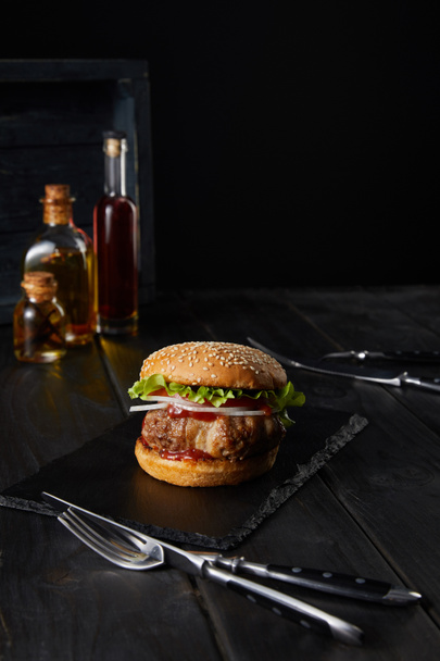 selective focus of burger on dark chopping board, fork, knife, oil and vinegar bottles isolated on black - Photo, Image