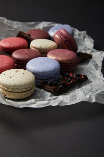 close up view of assorted delicious colorful french macaroons on crumpled paper with chocolate on black background - Fotoğraf, Görsel