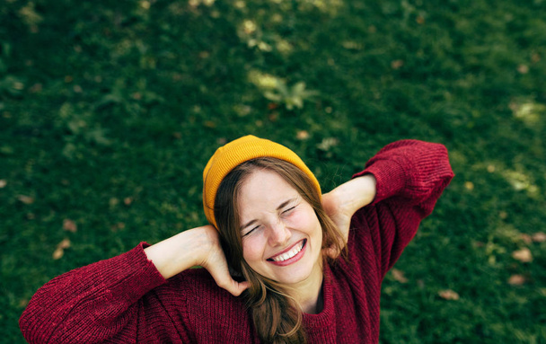 Above view image of a happy blonde young woman smiling broadly with a healthy toothy smile, wearing a red sweater, and yellow hat, posing on greenery nature background in the park. - Fotografie, Obrázek