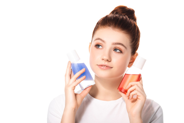 thoughtful teenager holding two bottles with red and blue makeup removers, isolated on white - 写真・画像