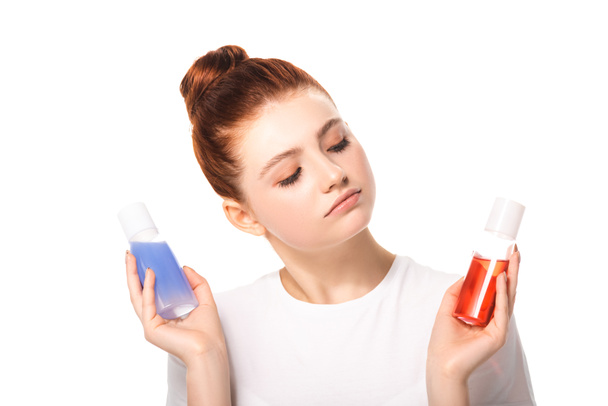 pensive teen girl holding two bottles with red and blue makeup removers, isolated on white - Фото, изображение