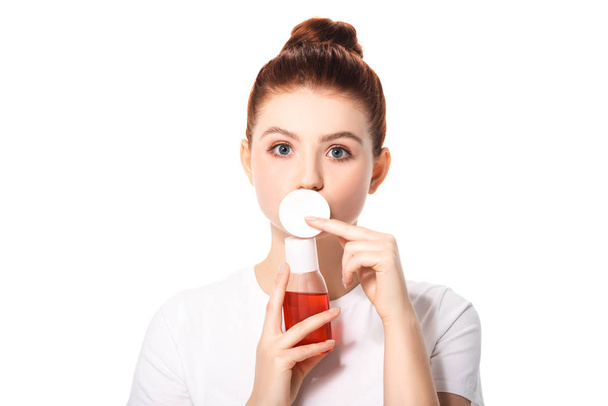 surprised teenager with clean skin holding cotton sponge and bottle with red makeup remover, isolated on white - Foto, Imagem