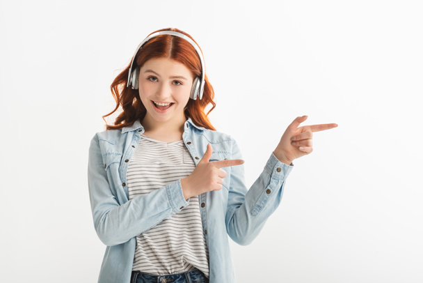 cheerful redhead teen girl listening music with headphones and pointing isolated on white - Valokuva, kuva