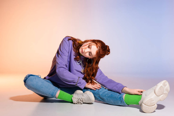 schöne gelangweilte Teenie-Mädchen mit roten Haaren sitzt auf lila und beige  - Foto, Bild