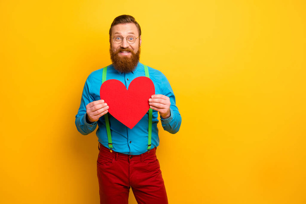 Photo of handsome guy hold hands big paper heart postcard call girlfriend romantic date wear blue shirt green suspenders red trousers isolated yellow color background - Foto, Imagem