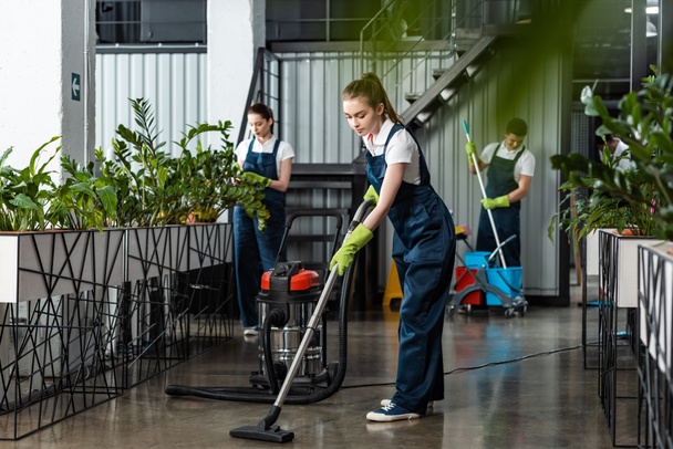 Genç temizlikçi ofis zeminini meslektaşlarının yanında süpürüyor - Fotoğraf, Görsel