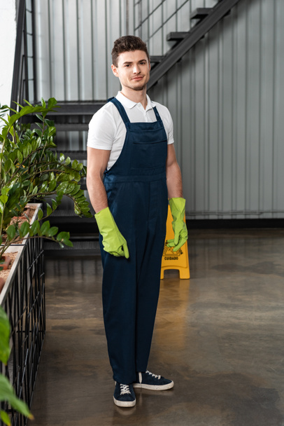 glimlachende jonge schoonmaakster in overall en rubberen handschoenen kijkend naar camera - Foto, afbeelding