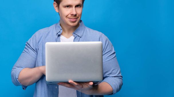Egy férfi alkalmi ruhában, kék háttérrel gépel egy laptopon, és a kamerába néz. Közelről.. - Fotó, kép