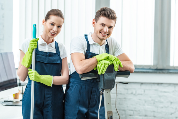 two young cleaners in overalls smiling at camera while standing in office - Foto, immagini