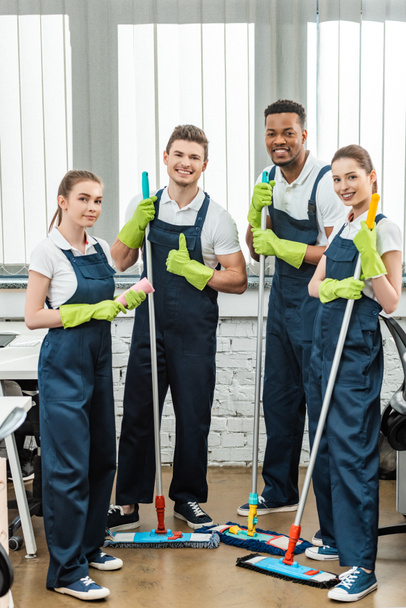 happy multicultural team of cleaners looking at camera and showing thumbs up while holding mops - Photo, Image