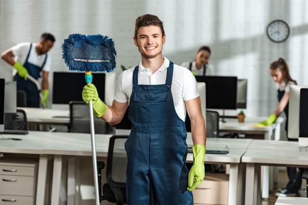 cheerful cleaner holding mop while looking at camera near multicultural colleagues - Photo, Image