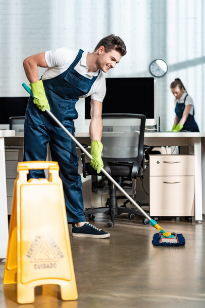smiling cleaner washing floor with mop near colleague cleaning desk - Photo, Image