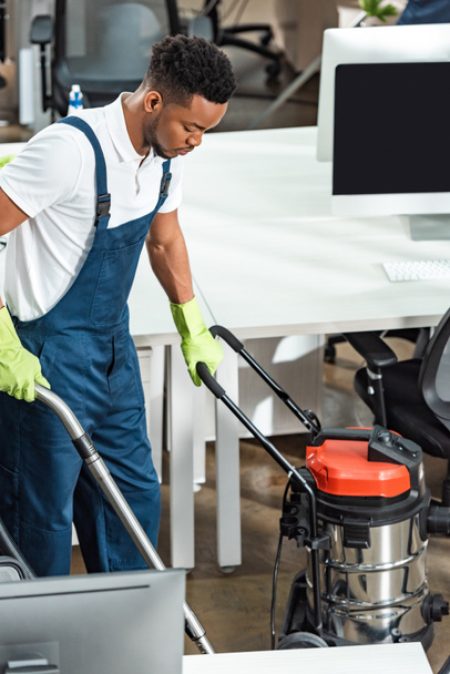 jovem afro-americano homem limpeza chão no escritório com aspirador de pó
 - Foto, Imagem