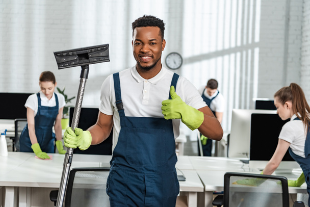 sorrindo afro-americano limpador segurando escova aspirador de pó e mostrando polegar perto da equipe de colegas
 - Foto, Imagem