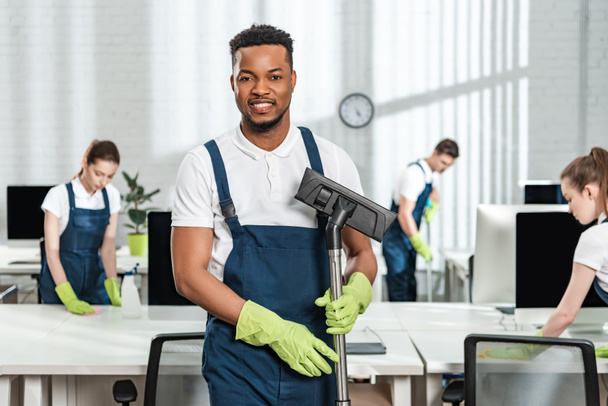 smiling african american cleaner holding vacuum cleaner brush near team of multicultural colleagues - Photo, Image