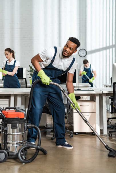 smiling african american cleaner vacuuming floor while looking away - Photo, Image