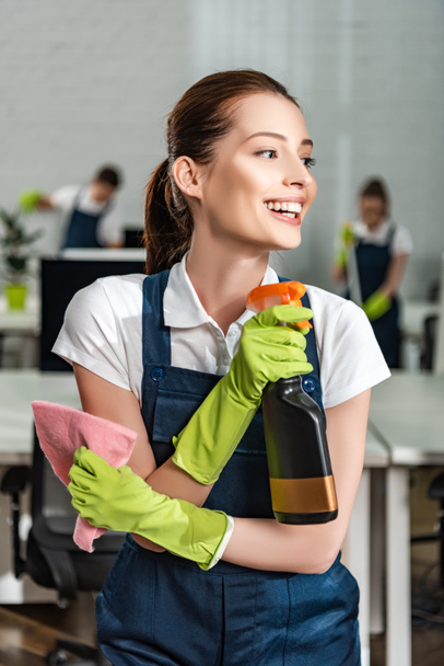 happy cleaner looking away while holding detergent spray and rag - Photo, Image