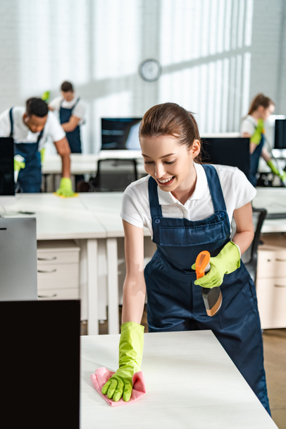 cheerful cleaner in overalls cleaning office desk with rag - Photo, Image