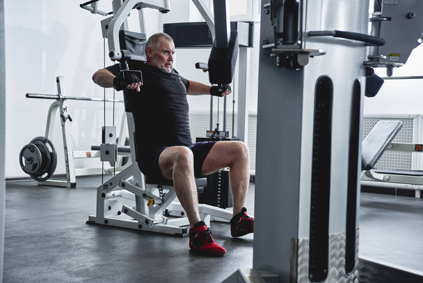 Adult gray-haired man trains on fitness equipment in the gym, pumps legs and arms muscles, loses weight. Concept of healthy lifestyle in old age - Valokuva, kuva