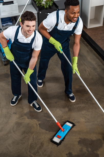 dois limpadores multiculturais sorridentes lavando o chão com esfregonas
 - Foto, Imagem