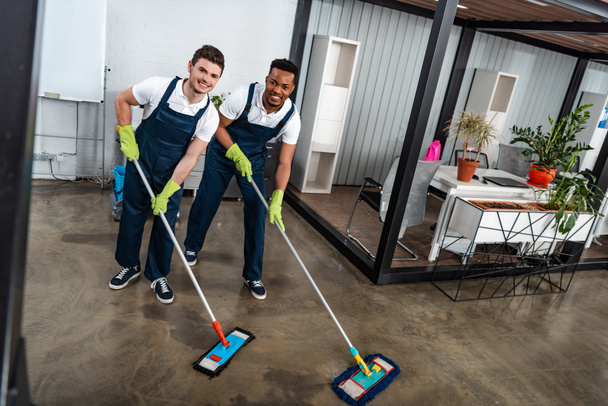 two smiling multicultural cleaners washing floor with mops and looking at camera - Photo, Image