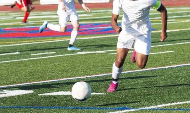 High school ragazzi partita di calcio con un atleta che insegue th eball giù per il campo indossando una divisa bianca
. - Foto, immagini