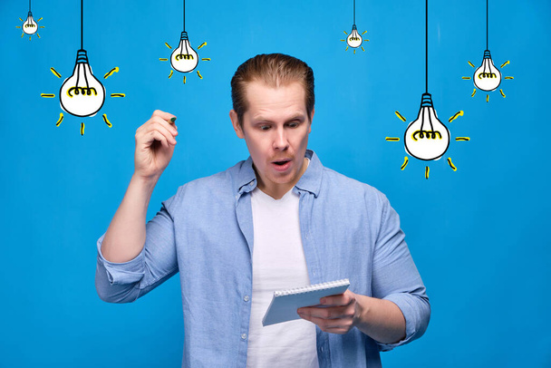 A man in casual clothes on a blue background holds a notebook and a pen wants to write down an idea. Abstract light bulbs burn on the background. Close up. - Fotoğraf, Görsel