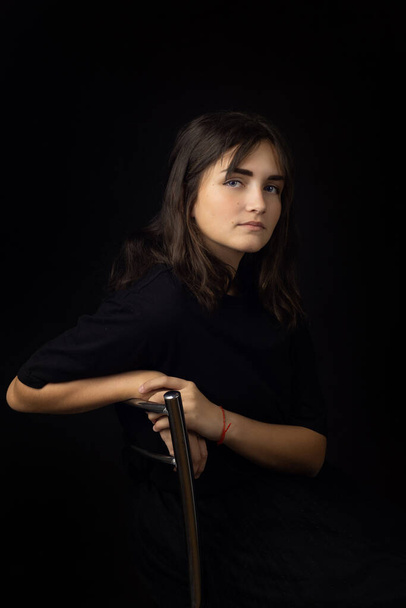 Πορτρέτο ενός δεκαπεντάχρονου κοριτσιού σε μαύρο φόντο, ένα κορίτσι θολωμένο στο πίσω μέρος μιας καρέκλας - Φωτογραφία, εικόνα