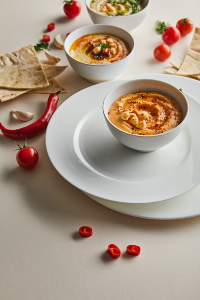 Selective focus of plates, bowls with hummus, cherry tomatoes, chili peppers, garlic and pita bread on grey  - Foto, Bild