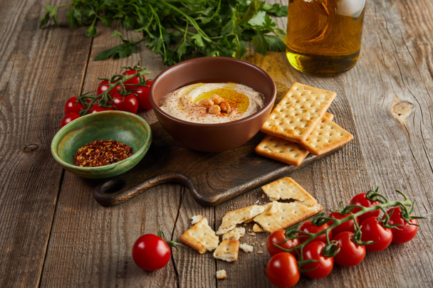 Crackers, bowls with hummus and spices on cutting board, vegetables and jar of olive oil on wooden background - Photo, Image