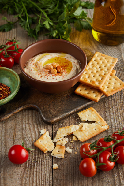 Crackers, bowls with hummus and spices on cutting board, vegetables, parsley and jar of olive oil on wooden background - Foto, immagini