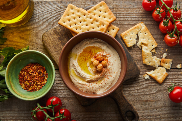 Draufsicht auf Cracker, Schalen mit Humus und Gewürzen, Gemüse, Petersilie und einem Glas Olivenöl auf Holzgrund - Foto, Bild