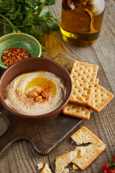 Selective focus of crackers, parsley, bowls with paprika and hummus on cutting board on wooden background - Фото, зображення