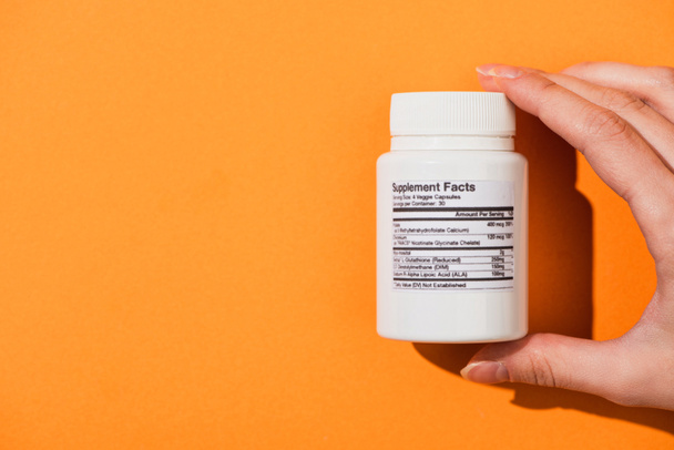 Vista parcial de la mujer sosteniendo contenedor blanco con suplementos dietéticos sobre fondo naranja
 - Foto, Imagen