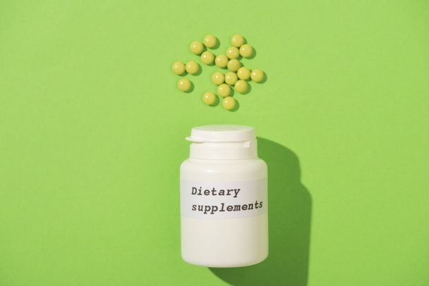 Top view of container with dietary supplements lettering and pills on green background - Photo, Image