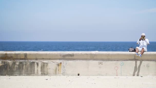 Az ember trombitál a tenger hátterén. Felvétel. Swarthy ember játszik trombita ül a töltésen a gyönyörű tenger. Zenészekkel való együttműködés a tengeri táj hátterében - Felvétel, videó