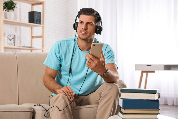 Man with mobile phone listening to audiobook on sofa indoors - Φωτογραφία, εικόνα