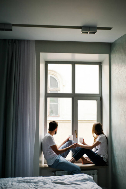 Man en vrouw bij het raam. Een regenachtige ochtend thuis. Jonge man en vrouw zitten op vensterbank met kopjes koffie. Isolatie, quarantaine.  - Foto, afbeelding