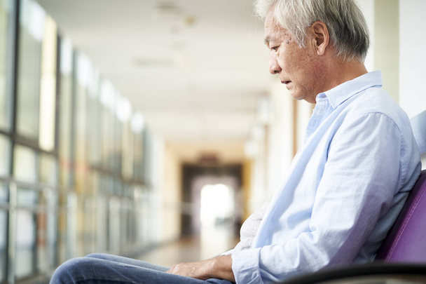 trauriger und verwüsteter asiatischer Senior sitzt allein in leerem Krankenhausflur, Kopf nach unten - Foto, Bild