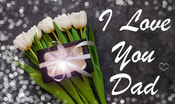 Μ 'αρέσει το σήμα του μπαμπά σου. Λευκά λουλούδια τουλίπας και μαύρο κουτί δώρου σε σκούρο φόντο με bokeh φώτα επίπεδη θέσει. Λουλούδι Μπουκέτο με ασημένια κορδέλα τόξο, συγχαρητήρια ευχετήρια κάρτα πάνω άποψη. - Φωτογραφία, εικόνα